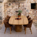 Patio dining setup featuring a teak oval outdoor dining table surrounded by six brown chairs in front of a stone wall
