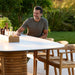 A man sits at a modern, oval outdoor dining table with white ceramic top surrounded by wooden chairs, with greenery in the background 