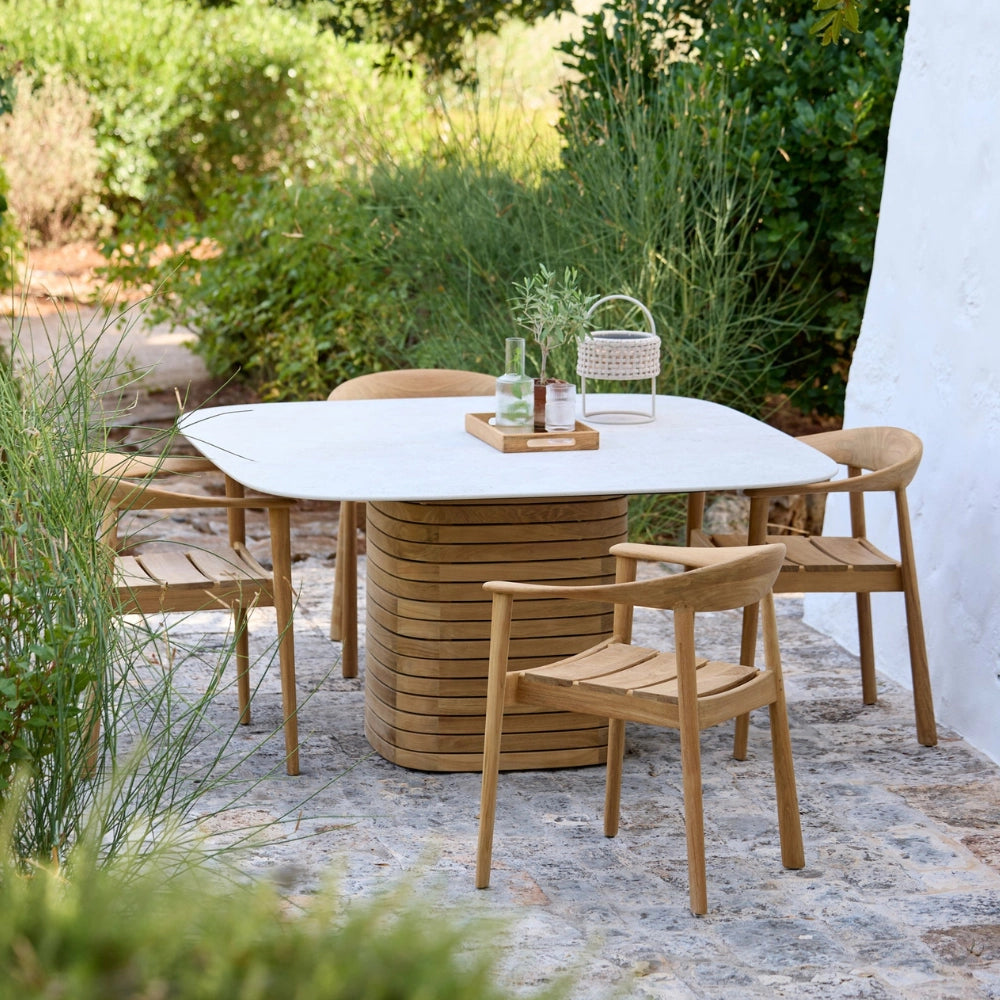 Square outdoor dining table with a wooden base and white ceramic top, paired with four wooden chairs on a patio surrounded by greener