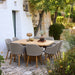Square wooden outdoor dining table with eight gray cushioned chairs on a patio, surrounded by greenery