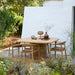 Patio scene featuring square wooden outdoor dining table and chairs surrounded with greenery