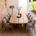 Oval teak outdoor dining table surrounded by teak chairs with gray cushions, set on a patio against a stone wall