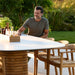 An oval teak outdoor dining table with a white tabletop, with a man sitting on a wooden chair on a patio surrounded by greenery