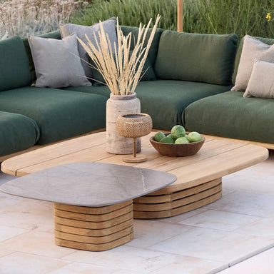 Patio lounge area with a green sectional sofa, a wooden square coffee table, and a small ceramic-top side table, with tall green grasses in the background