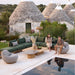 Outdoor seating area near a pool, with a woman sitting on a green sectional sofa, a man on a lounge chair, square wooden coffee tables, and traditional stone trulli in the background