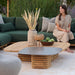 Patio lounge with a wooden square outdoor coffee table, small ceramic-top table, and a woman seated on a green sofa with greenery behind