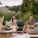 Patio seating area with a man and woman sitting, a large wooden outdoor coffee table and a small coffee table with gray ceramic top, surrounded by greenery