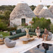 Outdoor seating area with green sectional sofa, wooden square coffee table, serene pool, and traditional stone trulli houses in the background