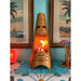 Gold cone shaped mini metal fire pit with tiki mask design with a flame inside, placed on a table with two tropical themed frames on the wall