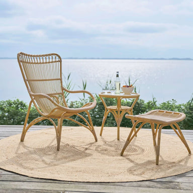 Natural rattan outdoor footstool with a matching lounge chair and side table on a seaside deck.