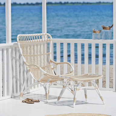White rattan outdoor high-back lounge chair beside a matching footstool on a seaside patio.