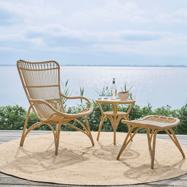 Natural rattan outdoor high-back lounge chair with a matching side table and footstool on a seaside deck.