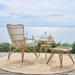 Natural rattan outdoor high-back lounge chair with a matching side table and footstool on a seaside deck.
