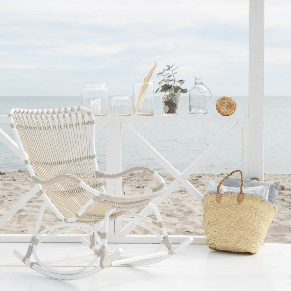 White rattan outdoor rocking chair on a seaside patio beside a woven storage basket.