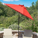 A bright red patio umbrella with a black pole stands on a stone patio in a garden setting, with two beige lounge chairs in the foreground and trees and blue sky behind.