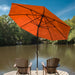 A large orange patio umbrella stands on a wooden dock over two brown Adirondack chairs, with a calm lake and trees in the background under a blue sky.