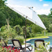 A large white patio umbrella with a brown metal pole stands over a poolside deck, shading lounge chairs in a landscaped backyard with trees, blue water, and a partly cloudy sky.