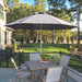 A large dark gray patio umbrella centered over a square outdoor dining table and four matching chairs on a stone patio, with a landscaped lawn and trees in the background.