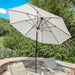 An off-white patio umbrella with a black pole and ribs, set up in an outdoor backyard patio environment with stone walls, lounge chairs, and green foliage.
