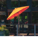 A large patio umbrella with a two-tone yellow and red canopy, mounted on a metal pole on an outdoor patio in front of a modern building with dark-framed glass windows.