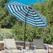 A blue and white striped patio umbrella is open on an outdoor stone patio, shading two cushioned chairs with a lantern in the center. Lush green garden foliage and pink flowers fill the background under a clear blue sky.