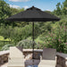 A large black patio umbrella stands over two beige outdoor lounge chairs on a stone patio, with a landscaped garden and trees in the background under a blue sky.