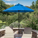 A bright blue patio umbrella with a black pole stands between two beige outdoor lounge chairs on a stone patio, with a lush green garden and trees in the background under a blue sky.