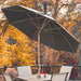 A large black patio umbrella with a beige pole and ribs is open on an outdoor deck, shading a wooden table and chairs. The background features a dense backdrop of bright orange and yellow autumn trees.
