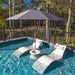 Outdoor poolside scene with two white lounge chairs, a round central table, and a large gray patio umbrella under a tree-lined deck railing.