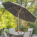 A large dark gray patio umbrella with a wooden pole and canopy ribs, shading a wooden dining table and four gray chairs on a deck with a lush green forest background.
