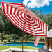 A large patio umbrella with bold red and white stripes stands open over a backyard pool. The scene includes green grass, trees, blue sky, and a small American flag hanging near the poolside furniture.
