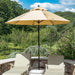 A large beige patio umbrella with a black pole stands between two matching beige lounge chairs on a stone patio, with a garden and blue sky in the background.