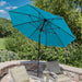 A bright turquoise blue patio umbrella with a black pole and canopy ribs, shading two outdoor lounge chairs on a stone patio with a garden background.
