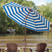 A large patio umbrella with bold blue and white stripes stands on a wooden deck overlooking a calm lake, with two brown Adirondack chairs in the foreground and trees in the background.