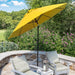 A bright yellow patio umbrella with a black pole stands over two outdoor lounge chairs on a stone patio, with green trees and pink flowers in the background.