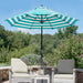 A large patio umbrella with teal and white stripes stands over a small table with a lantern, flanked by two outdoor lounge chairs on a stone patio with a garden background.