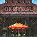 An outdoor patio scene in front of a brick building with a large red 'CENTRAL' sign. Bright orange patio umbrellas are set up under black-framed windows.