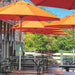 Bright orange patio umbrellas shading black picnic tables on an outdoor deck beside a brick building, with green trees and blue sky in the background.