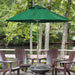 A large green patio umbrella stands on a wooden deck beside a fire pit table and four brown Adirondack chairs, with trees and water in the background.