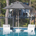 A large black patio umbrella stands over a swimming pool, with two white lounge chairs and gray pillows placed beside it. Trees and a house are visible in the background behind a dark railing.
