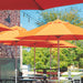 Bright orange patio umbrellas at an outdoor dining area with brick buildings, green trees, and string lights in the background.