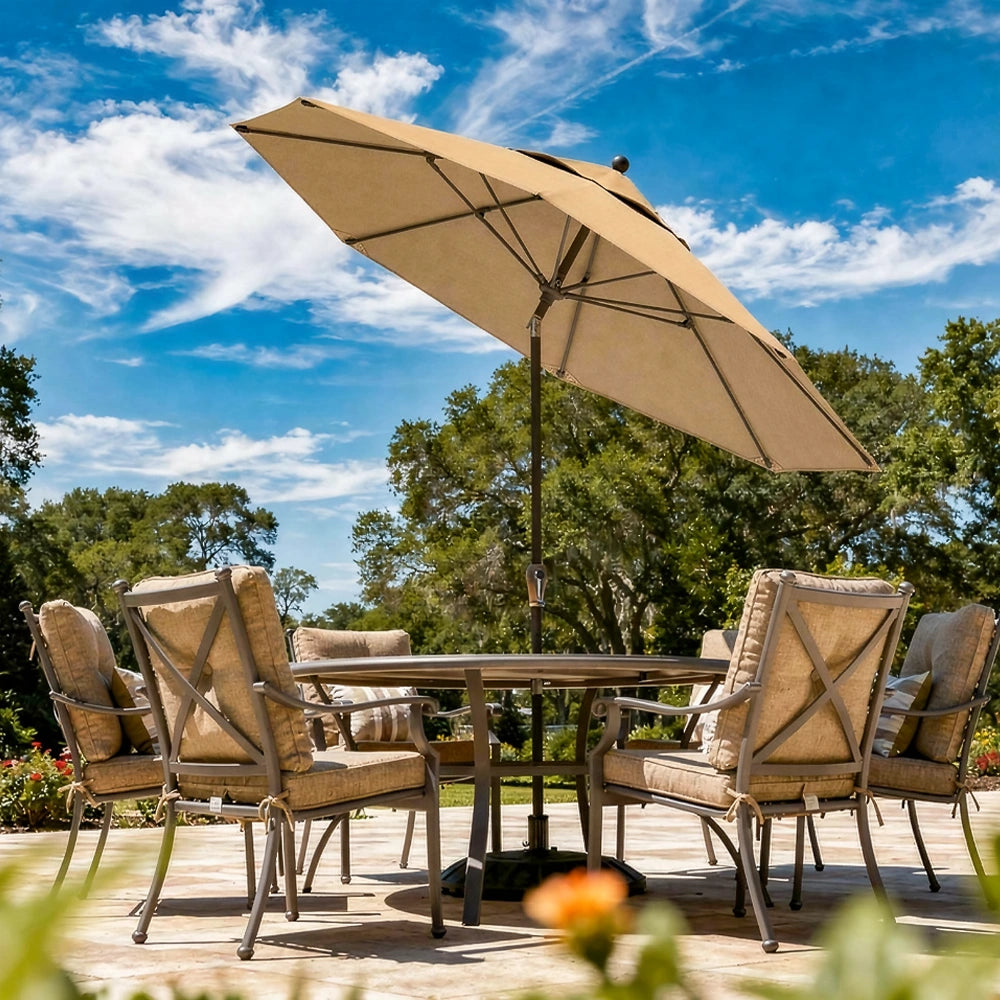 A beige patio umbrella stands over a round outdoor dining table with four cushioned armchairs on a patio, with trees and a blue sky in the background.