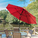 A large red patio umbrella with a black pole and ribs is open on a wooden deck beside a lake, shading a small round table and chairs. Trees and blue sky are visible in the background.