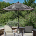 A large patio umbrella with bold black and white horizontal stripes stands on a stone patio between two cushioned outdoor chairs. The background features a lush green garden with trees and shrubs under a blue sky.
