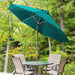 A teal patio umbrella stands open over a small round metal table with four beige patio chairs, set outdoors in front of a dense green forest background.