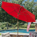 A large red patio umbrella with a brown metal frame is set up over a poolside outdoor seating area, with a swimming pool, trees, and an American flag visible in the background.