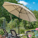 A large beige patio umbrella with a black pole is open in an outdoor garden setting, shading lounge chairs near a pool with blue sky and green trees in the background.
