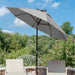 A large patio umbrella in a light gray color stands over two outdoor lounge chairs on a stone patio, with a lush green garden and blue sky in the background.