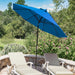 A bright blue patio umbrella with a black pole stands between two beige lounge chairs on a stone patio, with a lush green garden and trees in the background.