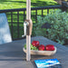An outdoor patio scene with a beige metal patio umbrella pole and crank handle inserted into a dark gray slatted table. A wooden bowl with red apples and green grapes sits on the table, with a blue magazine in the foreground and a garden background.